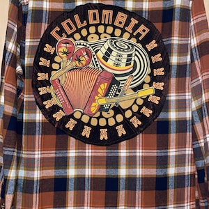 Könnte beinhalten: Braunes, marineblaues und weißes kariertes Flanellhemd mit einem großen runden Aufnäher auf dem Rücken. Der Aufnäher zeigt das Wort "COLOMBIA" und Illustrationen eines Hutes, eines Akkordeons und anderer kultureller Symbole.