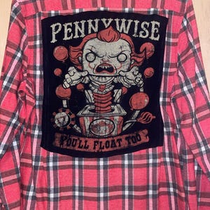 Può includere: Camicia di flanella a quadri rossi e neri con una grafica di Pennywise sul retro. La grafica presenta il testo "Pennywise" sopra un'immagine a cartoni animati del personaggio e il testo "You'll Float Too" sotto.