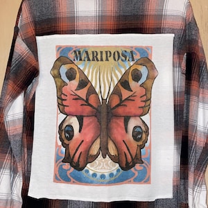 Könnte beinhalten: Ein rot-schwarz-weiß kariertes Flanellhemd mit einem Schmetterlingsmotiv auf der Rückseite. Das Motiv zeigt einen Schmetterling mit roten, orangefarbenen und braunen Flügeln und dem Wort "MARIPOSA" darüber.
