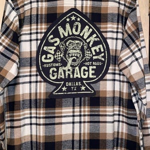Może przedstawiać: Koszula w kratę z flaneli w kolorze czarnym, białym i brązowym. Z tyłu duża naszywka z napisem "Gas Monkey Garage" i grafiką małpy. Koszula wisi na czarnym wieszaku.