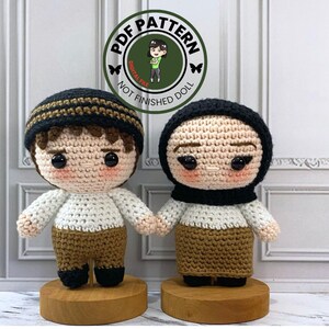 Puede incluir: Dos muñecas de crochet, una con un sombrero marrón y negro y la otra con un pañuelo negro. Ambas muñecas llevan pantalones marrones y camisas blancas. Las muñecas están de pie sobre bases de madera.