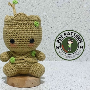 Könnte beinhalten: Gehäkeltes Amigurumi-Spielzeug eines grünen Charakters mit braunem Körper, grünen Blättern und schwarzen Augen. Das Spielzeug sitzt auf einem Holzsockel. Der Text "PDF PATTERN DIGITAL FILE NOT FINISHED DOLL" befindet sich auf einem grünen Kreis mit einem Schmetterling auf jeder Seite.