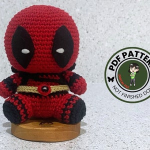 May include: Crocheted amigurumi Deadpool doll pattern. The pattern is a digital PDF file and does not include the finished doll. The doll is red and black with a white eye mask and a brown belt.