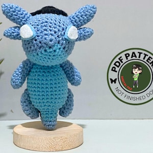 Könnte beinhalten: Handgefertigte blaue Häkeltierpuppe mit weißen Akzenten und einem schwarzen Kopfschmuck. Die Puppe steht auf einem kleinen Holzsockel. Ein runder Aufkleber zeigt "PDF PATTERN" mit einem Cartoon-Bild und dem Text "DIGITAL FILE NOT FINISHED DOLL".