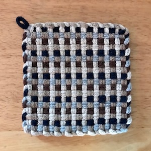 Op de afbeelding: Een geweven pot onderzetter met een geruit patroon in tinten bruin, blauw en beige. De pot onderzetter heeft een lus om op te hangen.