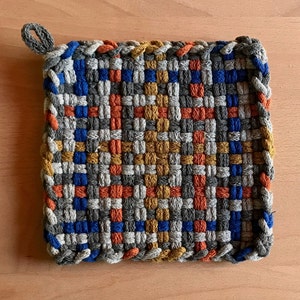 May include: A square woven pot holder with a grey, blue, orange, and gold pattern. The pot holder has a loop for hanging.