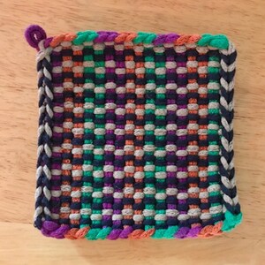 Könnte beinhalten: Ein quadratischer gewebter Topflappen mit einem bunten geflochtenen Rand in den Farben Orange, Grün, Lila und Grau. Die Mitte des Topflappens ist in einem karierten Muster mit den Farben Grau, Grün, Lila und Orange gewebt.