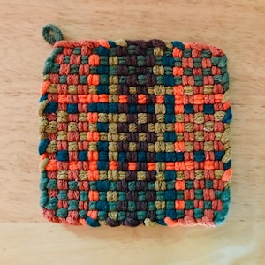 Puede incluir: Un posavasos cuadrado tejido con un asa de bucle. El posavasos está hecho con hilo naranja, marrón, verde y azul.
