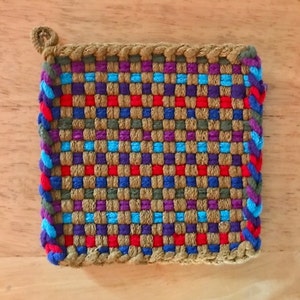 May include: A square woven pot holder with a brown, purple, blue and red checkered pattern. The pot holder has a brown braided border.