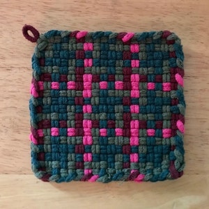 May include: A square woven pot holder with a loop handle. The pot holder is made with teal, dark green, and pink yarn.