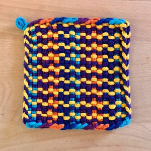 Könnte beinhalten: Ein bunter gewebter Topflappen mit einem blauen, gelben, orangen und violetten Muster. Der Topflappen hat einen geflochtenen Rand.