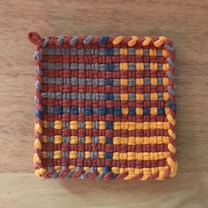 Può includere: Un sottopentola tessuto con forma quadrata e un bordo intrecciato. Il sottopentola è realizzato con filato arancione, giallo, blu e grigio.