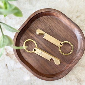 Könnte beinhalten: Zwei Flaschenöffner-Schlüsselanhänger aus Messing mit einem kreisförmigen Schlüsselring werden auf einem dunklen Holztablett präsentiert. Ein Schlüsselanhänger hat ein kleines geätztes Design. Die Schlüsselanhänger sind goldfarben.