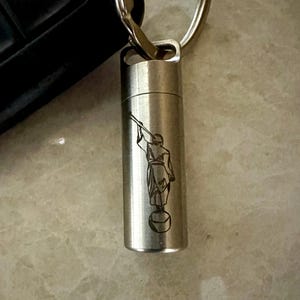 May include: A silver-colored keychain pendant with a cylindrical design. The pendant features an etched image of a person playing a horn. The keychain is attached to a silver ring.