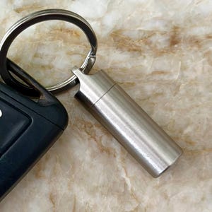 Op de afbeelding: Een zilverkleurige, cilindrische pillenhouder bevestigd aan een sleutelring, naast een autosleutelhanger. De pillenhouder heeft een schroefdop. De achtergrond is een lichtbruin en beige marmerpatroon.