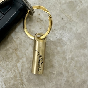 Peut inclure: Un porte-clés en métal doré avec un pendentif cylindrique. Le pendentif présente une image gravée en noir d'une personne soufflant dans une corne. Le porte-clés est attaché à un anneau doré, et une clé de voiture est visible en arrière-plan.