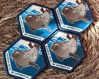 May include: Four hexagonal patches with a blue background and white border. Each patch features a cartoon image of a Saimaa ringed seal swimming in blue water. The text "Saimaa Seal of Approval" is printed in a black arc around the seal.