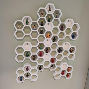 Può includere: Unità a scaffale a nido d'ape bianca con 28 scomparti esagonali, ognuno contenente una piccola statuina. Le statuette sono di vari colori e design, tra cui verde, rosso, blu e giallo.