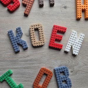 Può includere: Lettere colorate di mattoncini che compongono la parola "KOEN" su una superficie in legno chiaro. Le lettere sono rosse, blu, beige e bianche. Sono visibili anche altri mattoncini di vari colori.