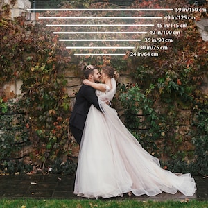 May include: A newly married couple embraces in front of a stone wall covered in autumn foliage. The bride wears a white wedding gown with a long train. The groom is in a black suit. Measurements are indicated in inches and centimeters.