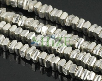 May include: Close-up of several strands of silver-colored, geometric beads. The beads have a slightly irregular, faceted shape and are strung together. The background is a dark, reflective surface.