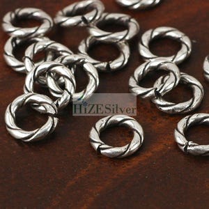 May include: A close-up of a group of silver-toned jump rings. The rings are twisted and have a smooth, polished finish. They are ideal for jewelry making and crafting.