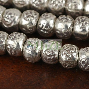 May include: Close-up of a strand of silver-colored beads. Each bead has a rounded shape with intricate embossed patterns. The beads are strung together, creating a textured and detailed appearance. The background is a dark brown surface.