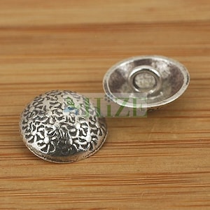 May include: Two silver-colored metal buttons with a textured, floral-like design on a wooden surface. One button is face up, and the other is face down, revealing the back. The buttons are likely for sewing or crafts.