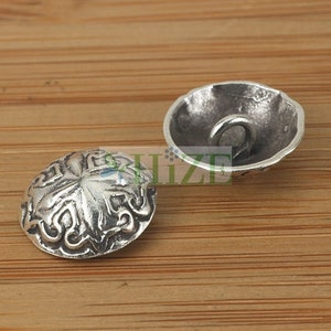 May include: Two silver metal buttons with an ornate design. One button has a raised design and the other is flat with a hole in the center.