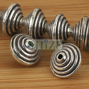 May include: Close-up of several silver-colored beads with a spiral design. The beads have a textured, antique finish and a hole for stringing. The beads are likely for jewelry making or other crafts.