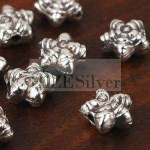 May include: Close-up of several silver-colored, flower-shaped beads. The beads have a detailed, textured surface and are scattered on a dark brown surface. The text "ZE Silver" is partially visible in the image.
