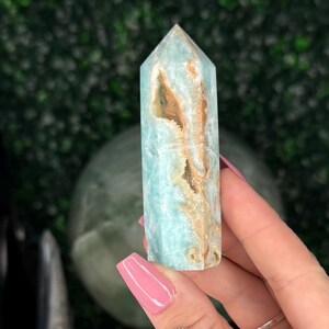 May include: A light blue and brown crystal tower is held in a hand with pink nail polish. Several other crystal towers are visible in the background, along with a green sphere and a black crystal.