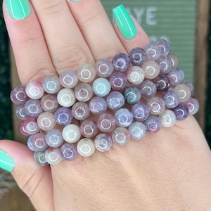 May include: A stack of purple, pink, and white gemstone bracelets. The bracelets are made of round beads and are stacked on a person's wrist.