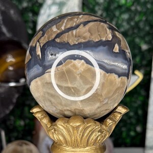 May include: A decorative sphere with a brown, white, and blue pattern, displayed on a gold-colored ornate stand. The stand has a detailed, vintage design. Other crystal spheres and points are visible in the background.