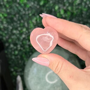 May include: A pink heart-shaped crystal is held up, showcasing its translucent interior. Several other pink heart-shaped crystals are displayed in a wooden tray. A black crystal tower and a green sphere are visible in the background, suggesting a theme of crystals and gemstones.