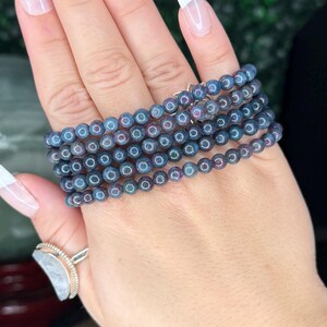 May include: A stack of beaded bracelets in shades of blue and purple, with a subtle iridescent sheen. The bracelets are displayed on a hand with a ring. The background features a green wall and decorative items.