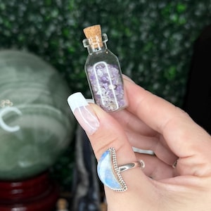 May include: A small glass bottle filled with purple and white contents, sealed with a cork. The bottle is held up against a green backdrop, with other similar bottles in the background. A silver ring with a blue stone is visible.