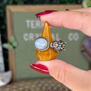 May include: A wooden ring display with a white grid pattern. The display has six pointed, orange crystal ring holders. A hand is holding a ring with a light blue stone on a pointed, orange crystal ring holder.