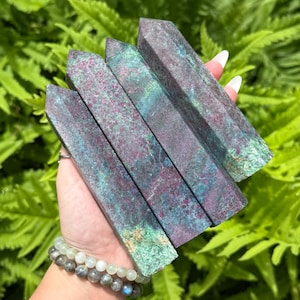 May include: Four rectangular crystals with a mottled teal and burgundy color are held in front of a backdrop of green foliage. The crystals have a polished, smooth surface. A beaded bracelet is visible on the wrist.