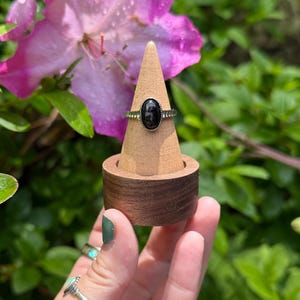 May include: A black oval gemstone ring on a wooden ring display stand. The ring is set in silver and has a delicate band.