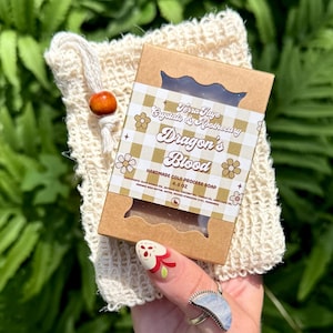 May include: A rectangular bar of Dragon's Blood handmade cold process soap in a brown cardboard box with a beige and white checkered label, placed inside a cream-colored woven fabric pouch with a small wooden bead on the drawstring closure.