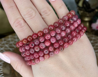 May include: A stack of pink and red gemstone bracelets with round beads. The bracelets are on a person's wrist.