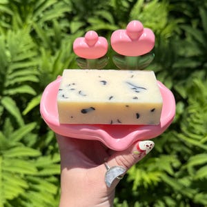 May include: A pink soap dish with a floral design holds a bar of soap. The soap is light-colored with dark flecks. Two pink flower-shaped accents are on the dish. The background is a lush green fern.