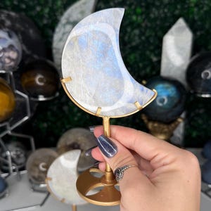 May include: A crescent moon-shaped crystal, with a gold-colored metal stand. The moon has a white and blue color scheme. The stand has a circular base and a thin stem. Other crystal spheres and objects are in the background.