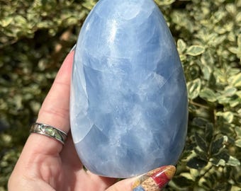 May include: A polished, egg-shaped blue calcite crystal with a smooth, glossy surface. The stone displays a translucent, pale sky-blue color with subtle white veining and internal crystalline structures visible throughout the mineral.