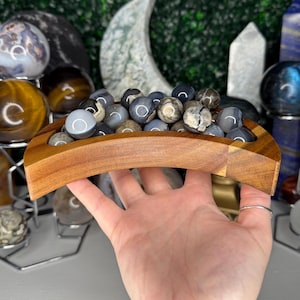 May include: A wooden bowl filled with gray and white agate spheres. The bowl is shaped like a crescent moon and is held in a person's hand.