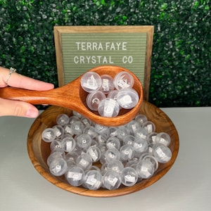 May include: A wooden bowl filled with clear plastic spheres containing small crystals. The spheres are being poured from a wooden spoon. A sign behind the bowl reads "Terra Faye Crystal Co."