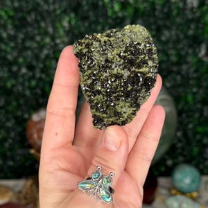 May include: A hand holds a rough, dark green and black mineral specimen. The mineral has a textured surface with small, sparkling crystals. A silver ring with a butterfly design is visible on the hand. Other crystals and the number "35" are in the background.