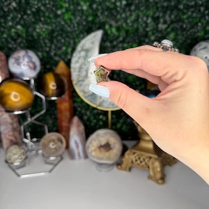 May include: A five-pointed star-shaped green and brown gemstone. The gemstone is being held between two fingers.