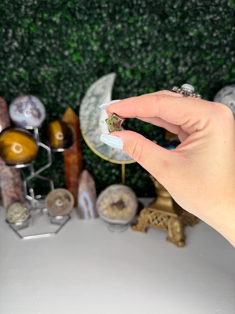 May include: A five-pointed star-shaped green and brown gemstone. The gemstone is being held between two fingers.